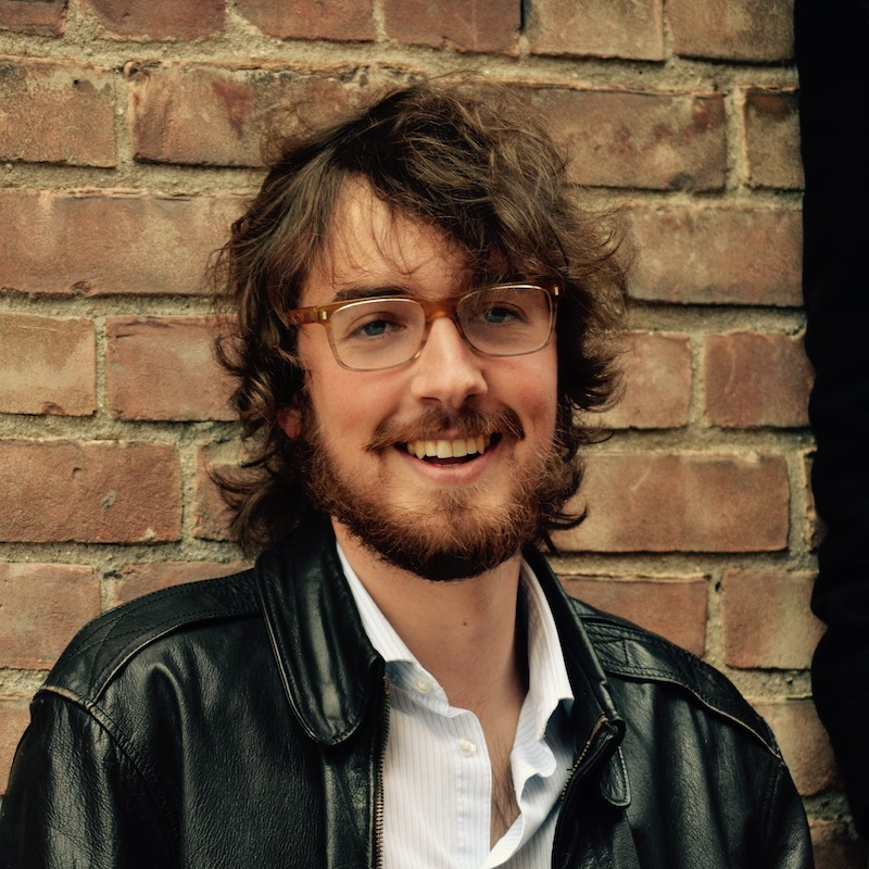 Jesse laughing, wearing leather jacket standing in front of brick wall